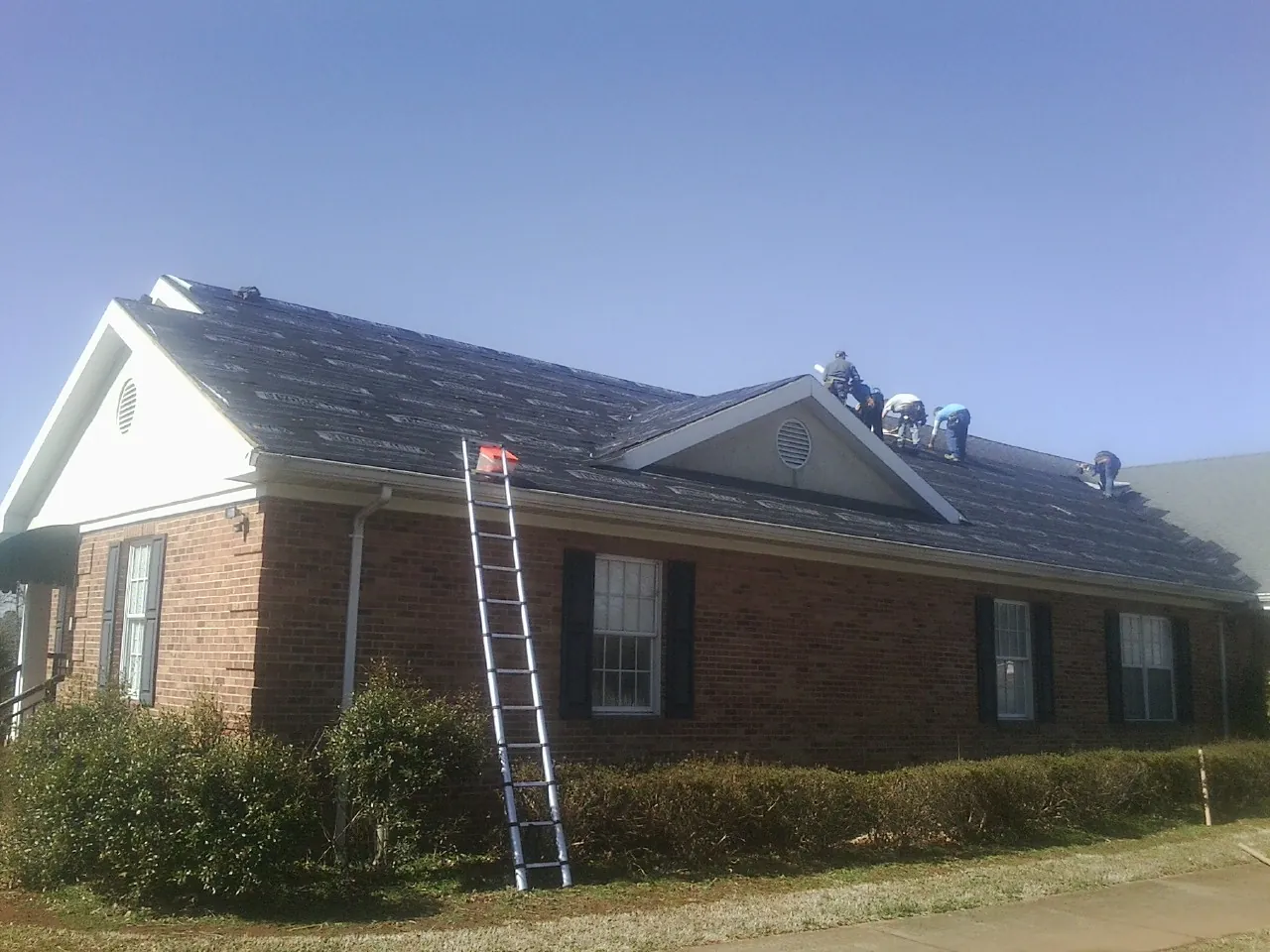 Crew tearing shingles off a brick ranch roof with a ladder set — Metal Roof Repair work by our crew in Whitten