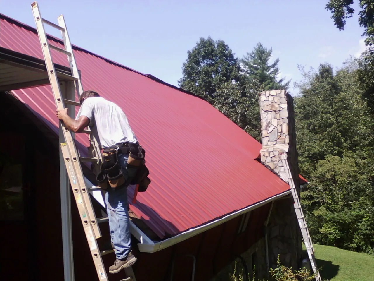 Roofer on a ladder beside a red metal roof and stone chimney — Soffit & Fascia Repair by Roofing Brothers in Whitten